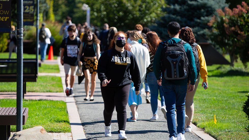 Students walking on the campus pathways wearing masks and Adelphi University sweatshirts.