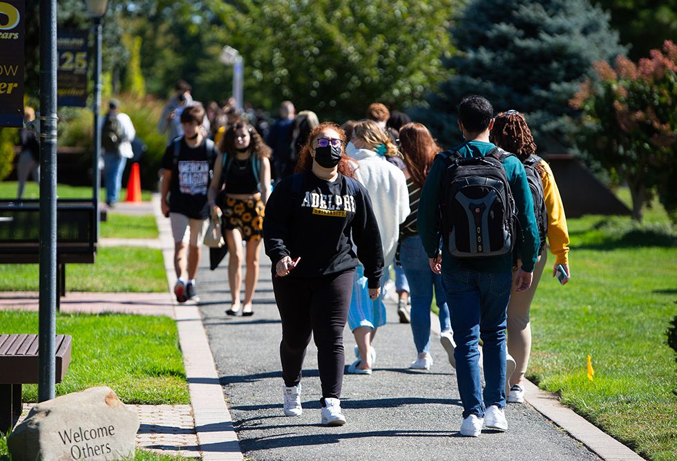 Students walking on the campus pathways wearing masks and Adelphi University sweatshirts.