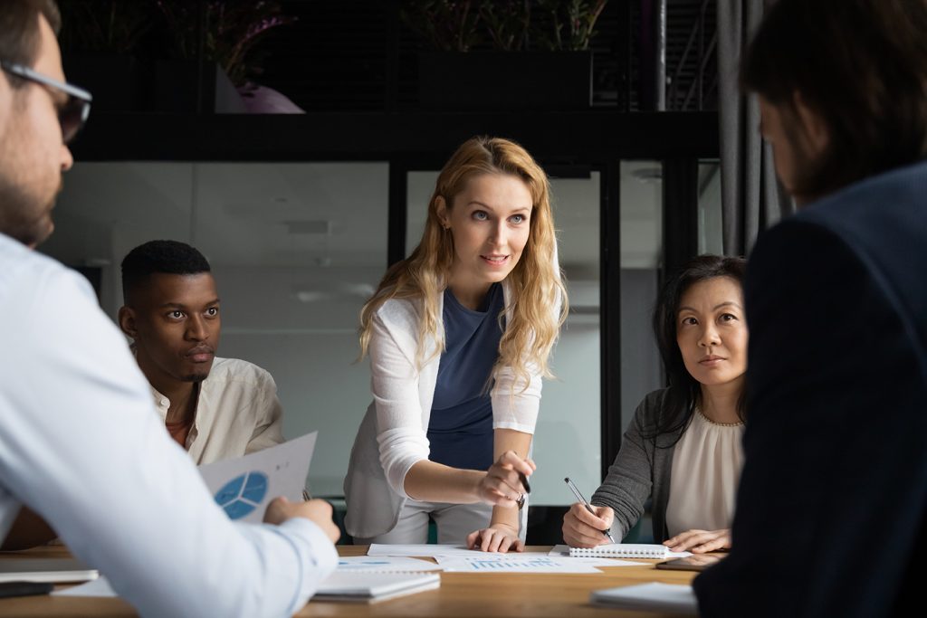 Confident skilled businesswoman explain marketing data paper report financial results analysis to multi-ethnic colleagues, company staff gathered at meeting brainstorming thinking strategizing concept