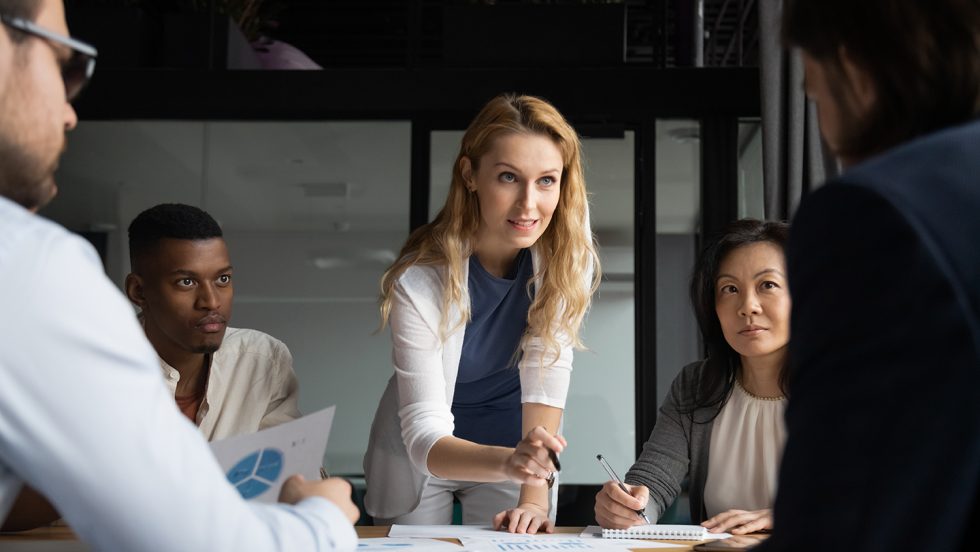 A group of professionals at a table. Female leader leans across while speaking.