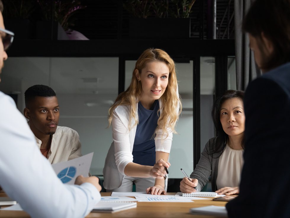 A group of professionals at a table. Female leader leans across while speaking.