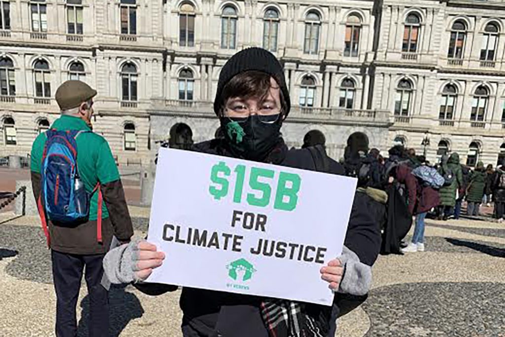 Student Kelly Andreuzzi is standing in front of capital in Albany holding a sign that reads Fifteen billion dollars for climate justice