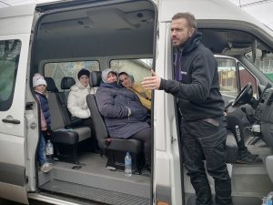 A man stands in the doorway of a bus filled with women and children in coats and hats.