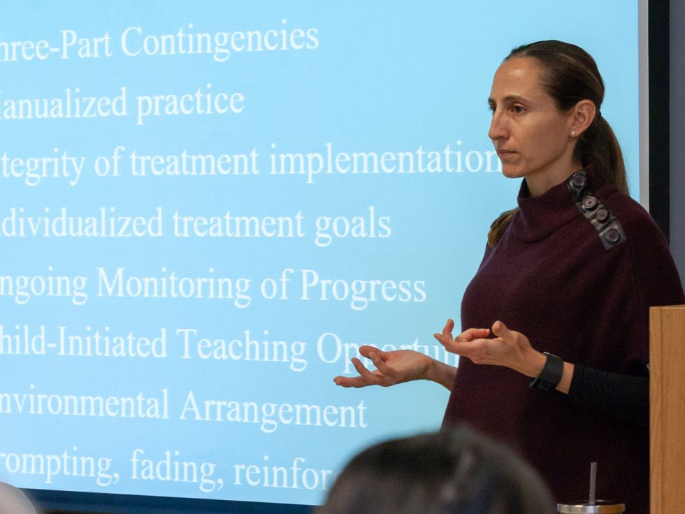 Dana Battaglia, PhD, standing in front of a slide presentation during a lecture.