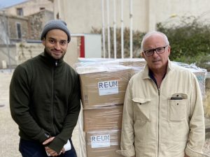 Two men stand in front of boxes marked REUM, which stands for Rescue & Equip Ukraine Mission