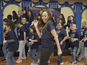 A woman in the foreground in a dance move, leading a group of elementary school children who are following her moves. The children's shirts have the words "PS 14Q Chorus." The curtain behind them reads "Fairview School Chorus."  