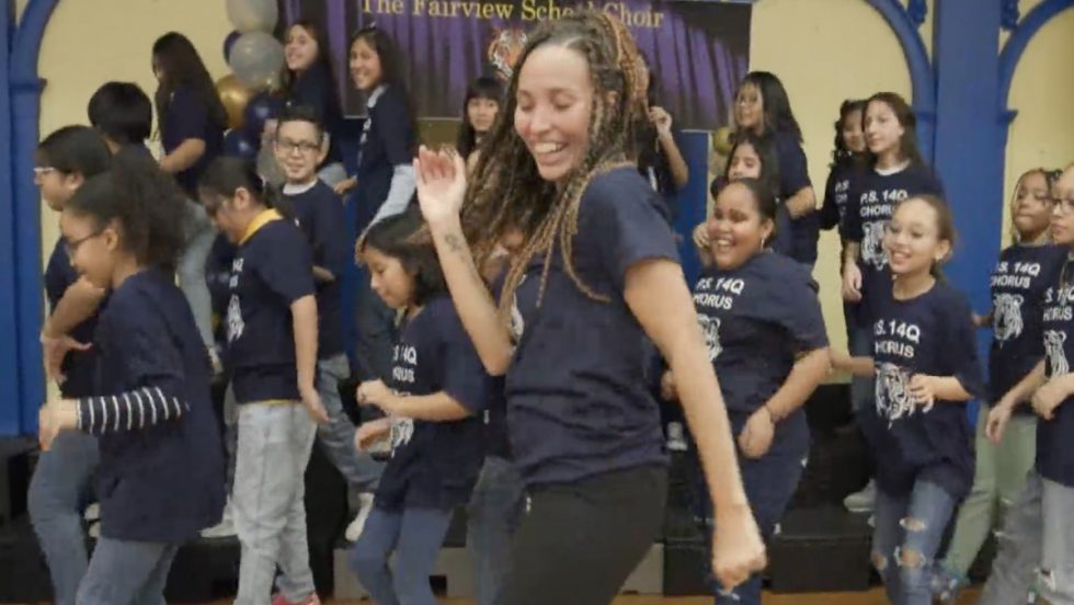 A woman in the foreground in a dance move, leading a group of elementary school children who are following her moves. The children's shirts have the words "PS 14Q Chorus." The curtain behind them reads "Fairview School Chorus."  
