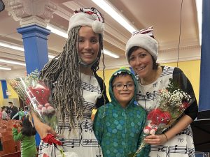 Two women wearing dresses with musical notes and Santa caps are smiling and holding bouquets of flowers. Standing between them is a smiling child in a green jacket.