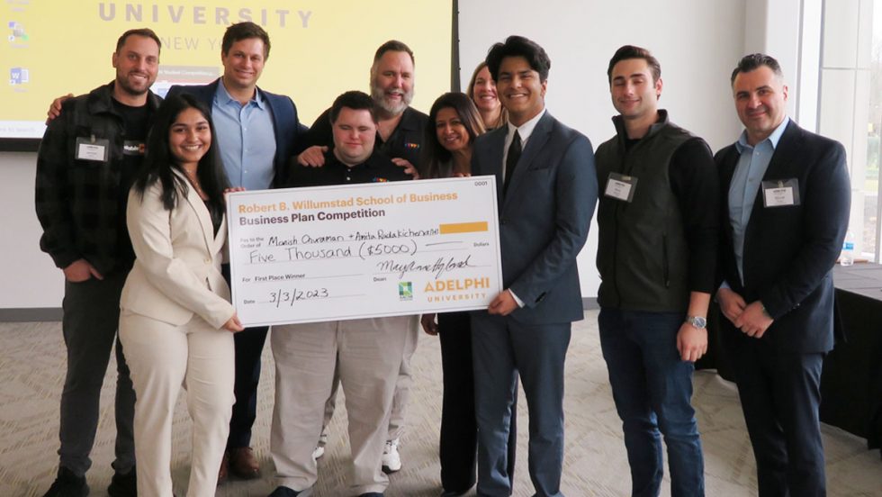 Two students, a woman in a beige pantsuit and a man in a dark suit, are holding a large check that reads: Robert B. Willumstad School of Business Business Plan Competition. Pay to the order of Monish Churaman and Amita Radakichenane, five thousand dollars. Signed by MaryAnne Hyland. Dated March 3, 2023. Adelphi University. Behind them stand eight people. In the background is a screen that reads Adelphi University New York.