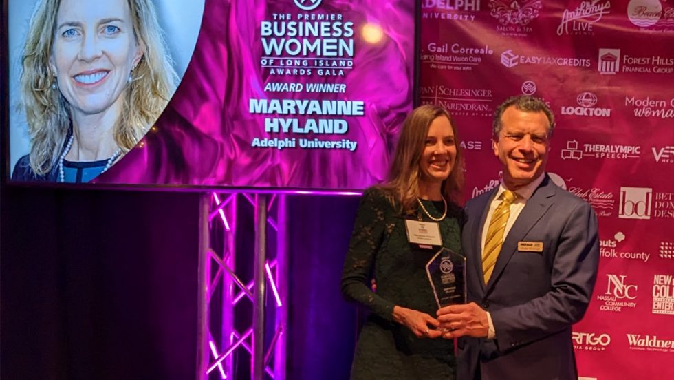 In foreground, MaryAnne Hyland is receiving her award from Stuart Richner. On the left is a presentation screen of her photo and the words "The Premier Business Women of Long Island Awards Gala, Award Winner MaryAnne Hyland Adelphi University." On the right is a backdrop with the names of all the organizations of the awardees, including Adelphi University.