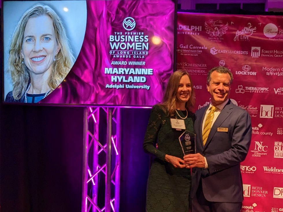 In foreground, MaryAnne Hyland is receiving her award from Stuart Richner. On the left is a presentation screen of her photo and the words "The Premier Business Women of Long Island Awards Gala, Award Winner MaryAnne Hyland Adelphi University." On the right is a backdrop with the names of all the organizations of the awardees, including Adelphi University.