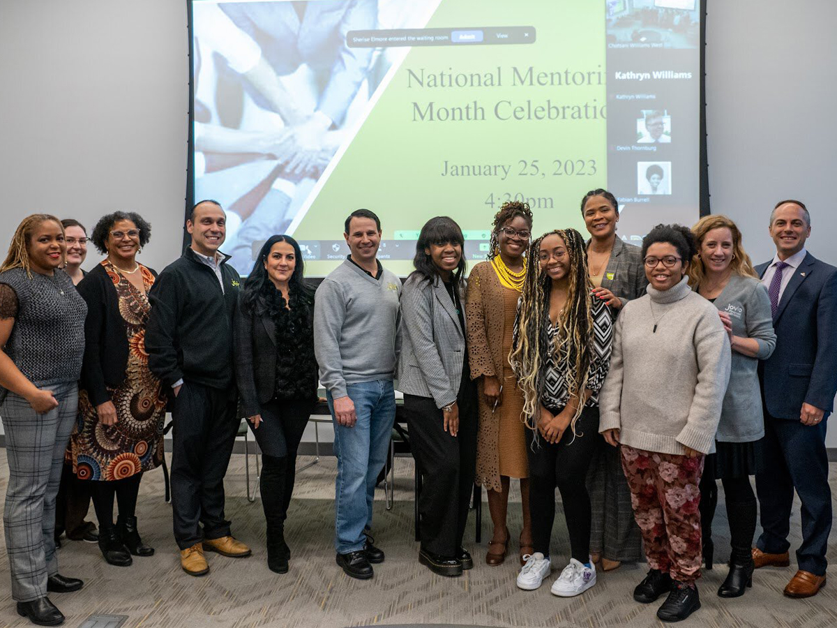 Students and mentors are standing in front of a screen that reads "National Mentoring Month Celebration, January 25, 2023, 4:30 p.m." To the left is a microphone at a podium with the words "Adelphi University