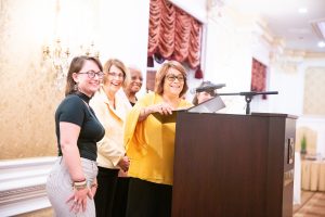 A white woman, smiling, with glasses, brown hair and a gold, loose top, stands in front of a podium. Behind her stand two white women wearing glasses, a Black woman with white hair and a white woman wearing a wide-brimmed black hat. They are all smiling.