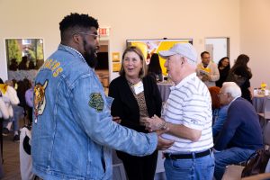 A middle-aged Black man in a denim jacket is in profile, smiling and talking with an older white man wearing a striped shirt, jeans and a baseball cap. A middle-aged woman with a brown bob wearing a brown blazer stands between and slightly behind them, looking on.