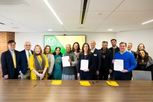 About 15 men and women of various ages and backgrounds stand behind a table for a group photo. They’re looking at the camera and smiling.