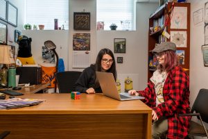 A white woman with long dark hair and glasses sits in an office at a desk. She is looking at a laptop screen to her left. A white female student with reddish-brown shoulder-length hair is typing on the laptop. She wears a red and black checked jacket, a shirt with a ruffle at the neck, dark-rimmed glasses and a houndstooth-print newsboy cap.