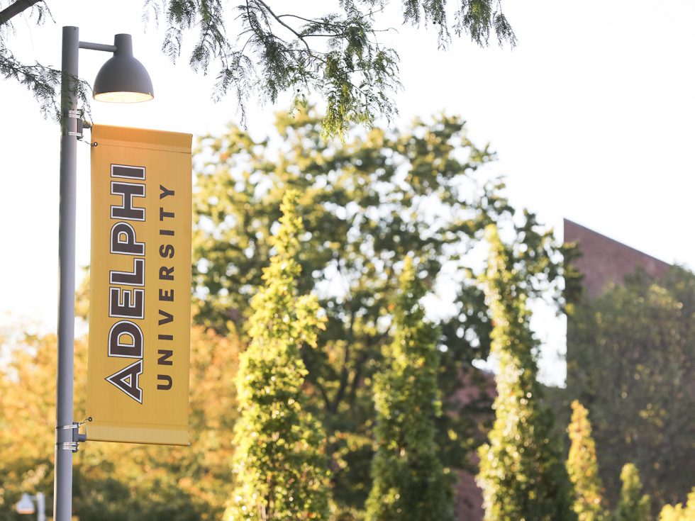 Adelphi University's main campus is located in Garden City, New York. Showing a gold sign with the Adelphi University logo and University Center in background.