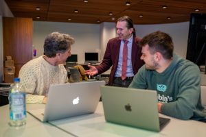 Two young white men in front of their laptops are turned away from the camera looking at their professor, a Hispanic man in a brown suit with a red tie, who is talking to them.