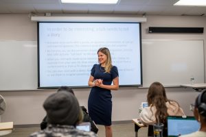 Woman in navy blue dress with blonde hair stands in front of classroom. Behind her is a projected screen saying, “In order to be interesting, a talk needs to tell a story.”