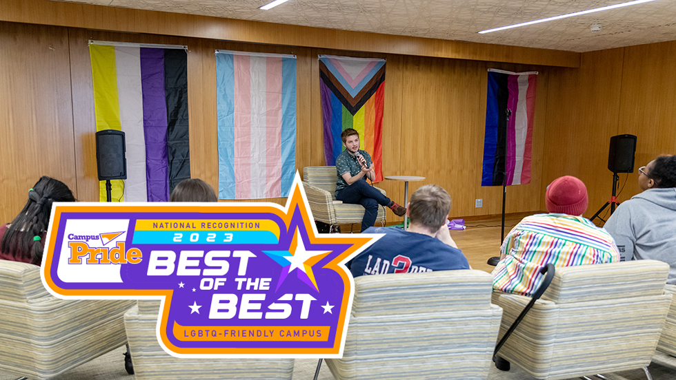 The Campus Pride Best of the Best Badge is displayed over a photograph of students in the Multicultural Center. The badge, which includes a large star as well as four smaller ones, also reads "National Recognition 2023" and "LGBTQ-friendly institution." In the photograph, a student holds a microphone and addresses a group of students, four of whom are seen. Four colorful LGBTQ and trans flags are on the wall behind the speaker.