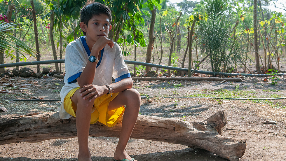A lonely young boy wearing shorts, a loose t-shirt and flip-flops, sits on a log in a Central American landscape.