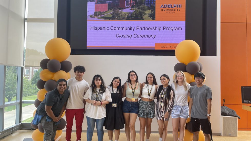 Nine students, male and female, of Hispanic descent, stand in front of a screen with the words “Hispanic Community Partnership Program Closing Ceremony: Adelphi University” and a photo of the Adelphi University campus. To their left and right are brown and gold balloons.