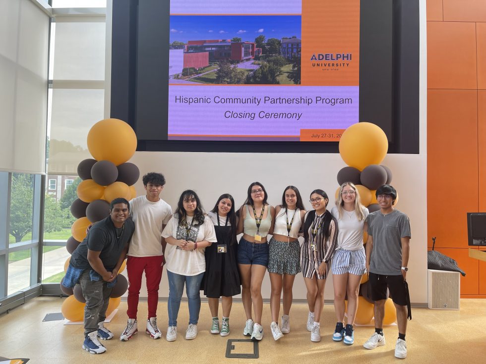 Nine students, male and female, of Hispanic descent, stand in front of a screen with the words “Hispanic Community Partnership Program Closing Ceremony: Adelphi University” and a photo of the Adelphi University campus. To their left and right are brown and gold balloons.