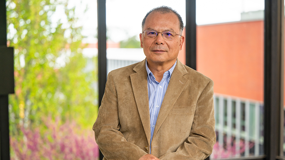  Dr. Huang facing the camera, standing in front of windows overlooking Adelphi's campus.