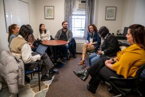 A white woman with brown curly hair wearing a blazer, white blouse, blue scarf and brown pants sits in a room. There are doctoral students, male and female, sitting on either side of her.