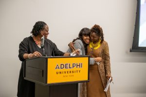 Black woman with brown hair and gold earrings wearing a brown dress stands at a podium with the words Adelphi University. A young Black woman wearing a gray blazer is on her left, smiling and hugging a Black woman with braids, also smiling, wearing a brown jacket with a yellow collar and brown skirt.