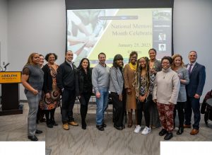 Thirteen people, male and female, of various ages and backgrounds, stand in front of a screen that reads National Mentoring Month Celebration: Adelphi University.