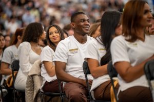 A young Black man is sitting in an audience with people seated in front of and behind him. They wear Adelphi University College of Nursing and Public Health scrubs.