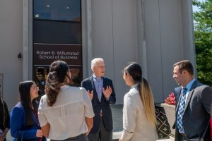 A white man with white hair and glasses wearing a suit is talking to students in front of a building. On the building is the name: Robert B. Willumstad School of Business.