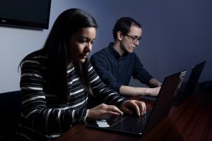 Two people on laptops, woman closest to camera, man furthest from camera. TV behind them.