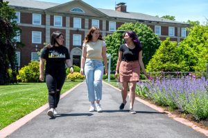 Three white young women walk on a path in front of Adelphi’s Levermore Hall. There are purple flowers growing on the right.