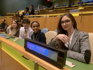 Four students in lecture hall in the United Nations: A white woman with brown hair and glasses is in the foreground. To her right sit a Black woman, a white man and a white woman.