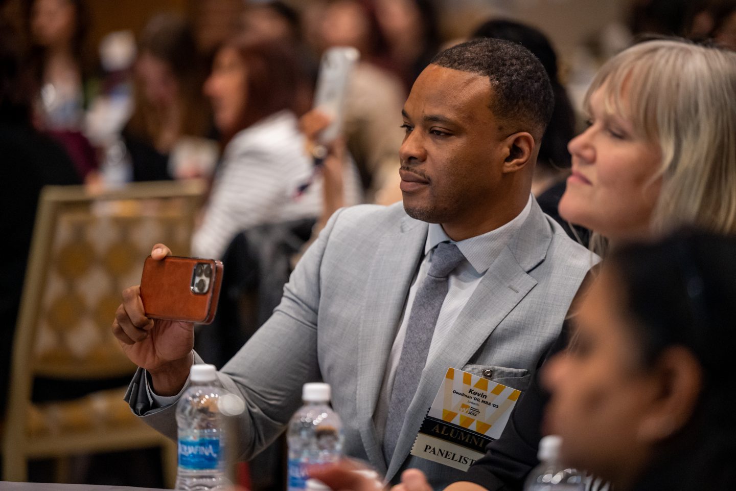 A Black man in a gray suit and tie is holding up a red phone to take a picture. Next to him, also looking to the left, is a white woman with blond hair.