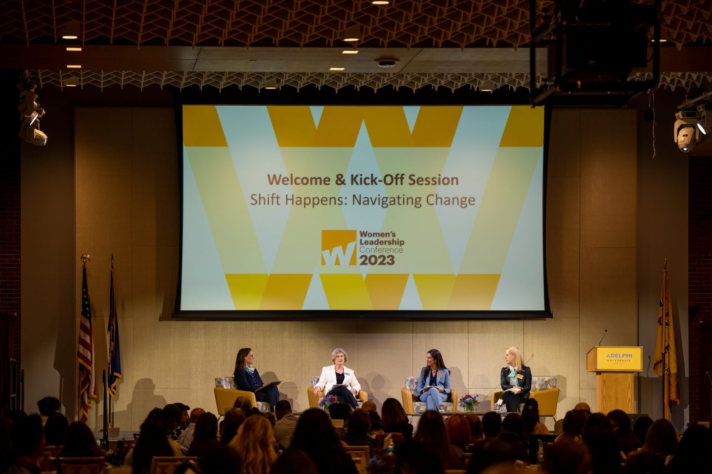 Four women are seated on chairs on a stage. The screen overhead reads: Welcome and Kick-Off Session, Shift Happens: Navigating Change, Women's Leadership Conference 2023. The sign on the podium reads: Adelphi University