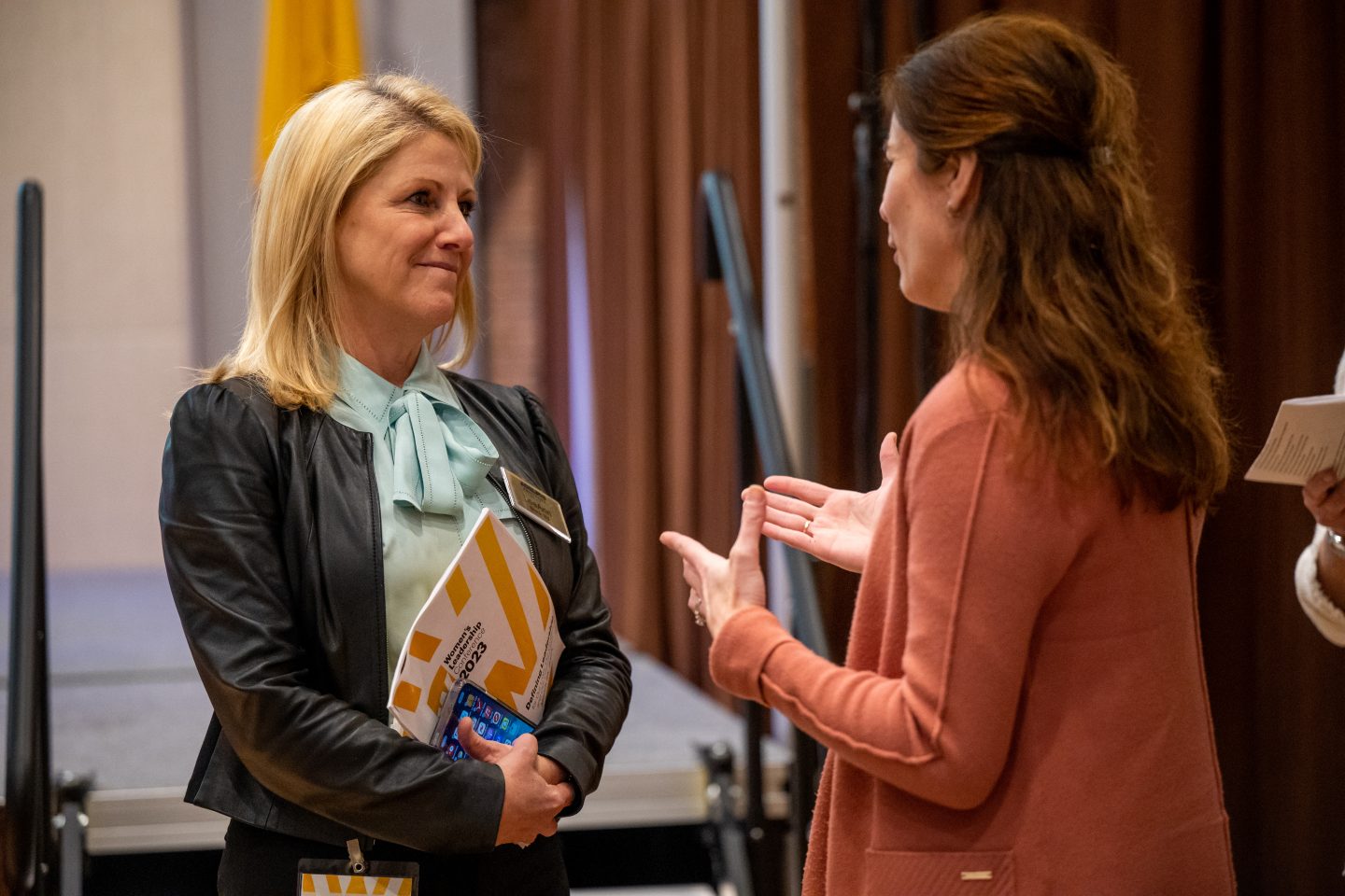 A woman with medium-length blond hair and bangs wearing a green blouse with a bow and green sweater is looking to the right at a woman with long brown hair and a brown shirt, who is holding out her hands as she speaks.