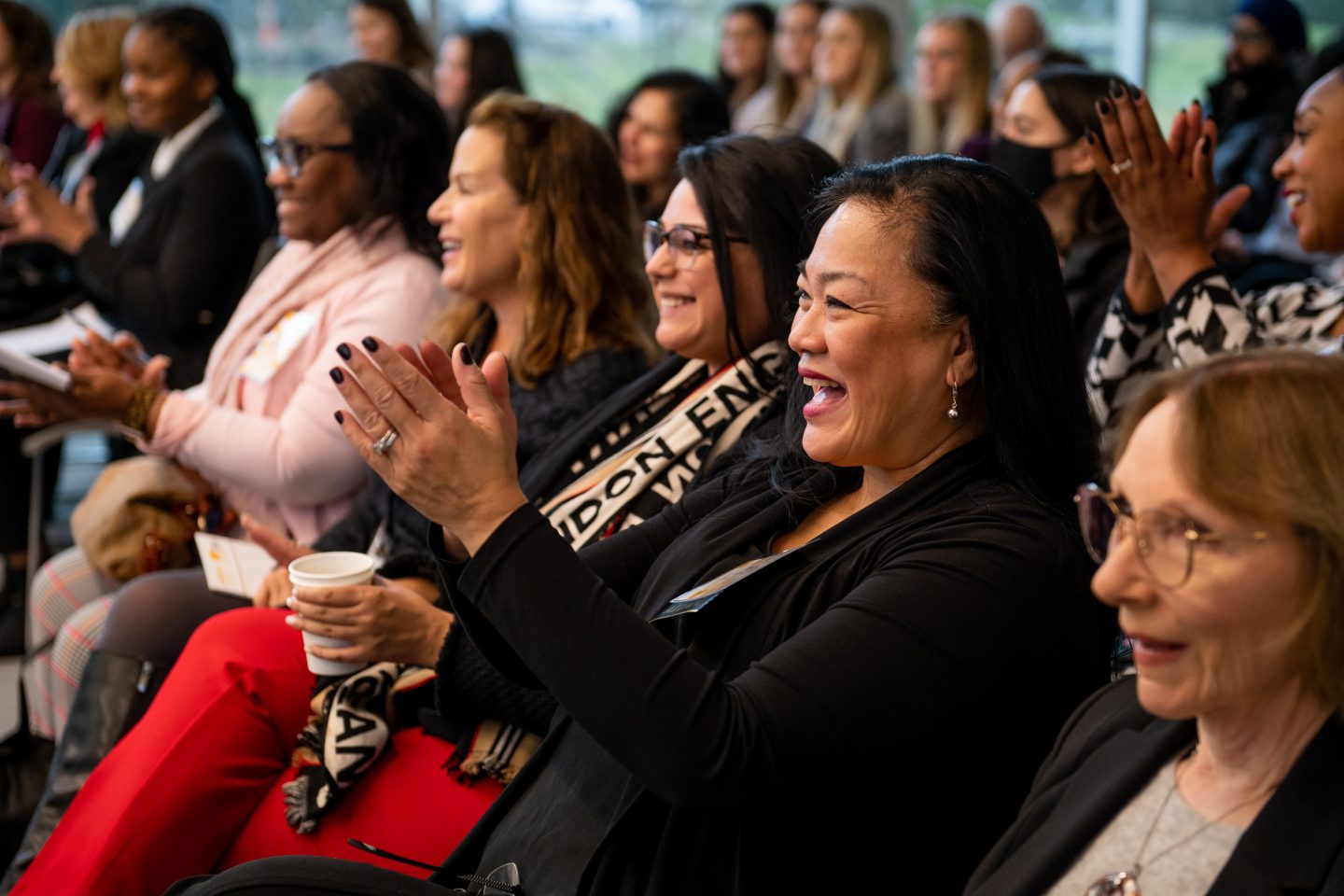 Four women in profile, seated in an audience, are smiling and applauding.