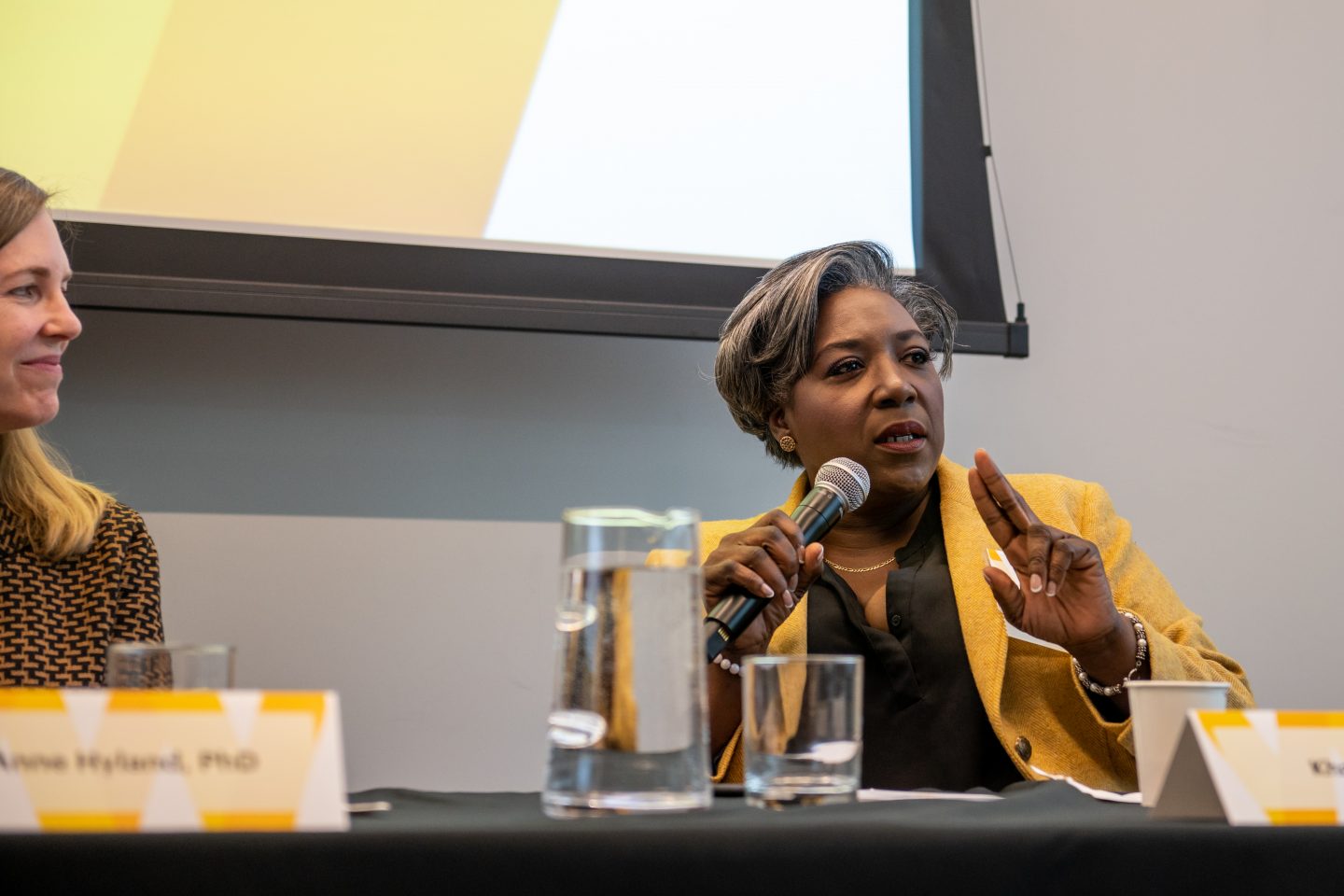 A Black woman with short grayish hair wearing a gold blazer is seated behind a table, holding a microphone and speaking while she gestures with her left hand.