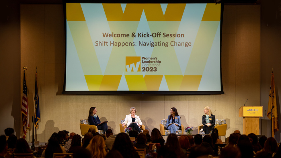 Four women are seated on chairs on a stage. The screen overhead reads: Welcome and Kick-Off Session, Shift Happens: Navigating Change, Women's Leadership Conference 2023. The sign on the podium reads: Adelphi University.