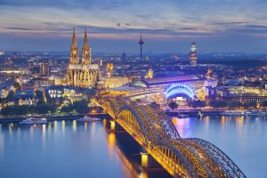 The city of Cologne at night.The city is lit up. There are two towers in the background and a bridge over water in the foreground.