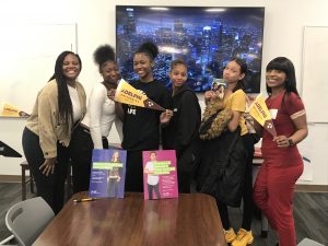 Four Black female students stand in front of a table displaying two books. They hold brown and gold pennants that read “Adelphi University.”