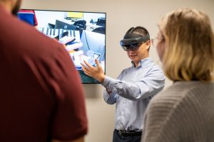 A man wearing virtual reality glasses holds his hand in front of him while a man and woman, with their backs to the camera, watch. They stand in front of an abstract painting