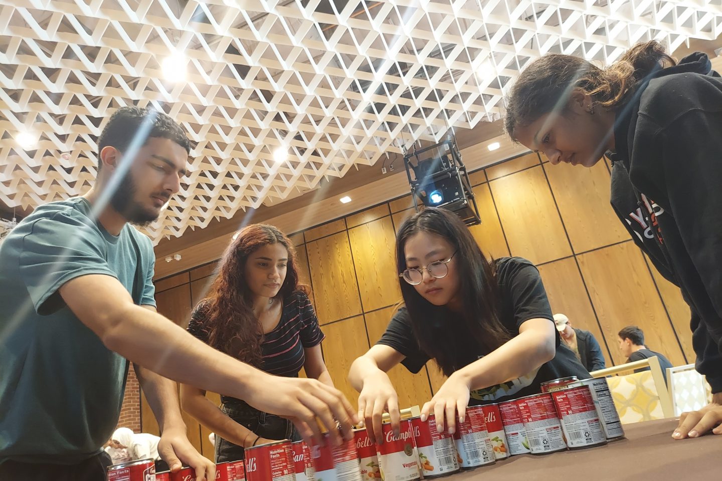 Four students leaning over table organize and arrange Campbell's soup cans.