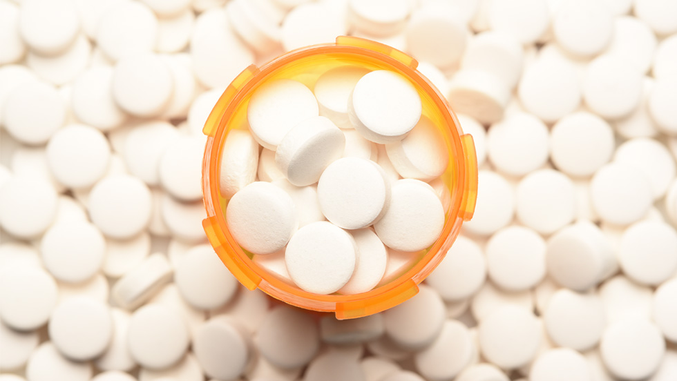 A photo of prescription drugs in a pill bottle. The bottle, seen from above, sits on a table completely covered with the same pills.