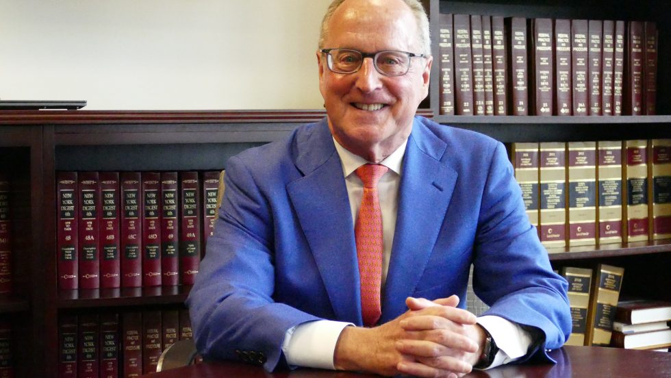 Bern Clair is wearing a blue suit, white shirt and an orange tie. He is sitting at a table and smiling into the camera.