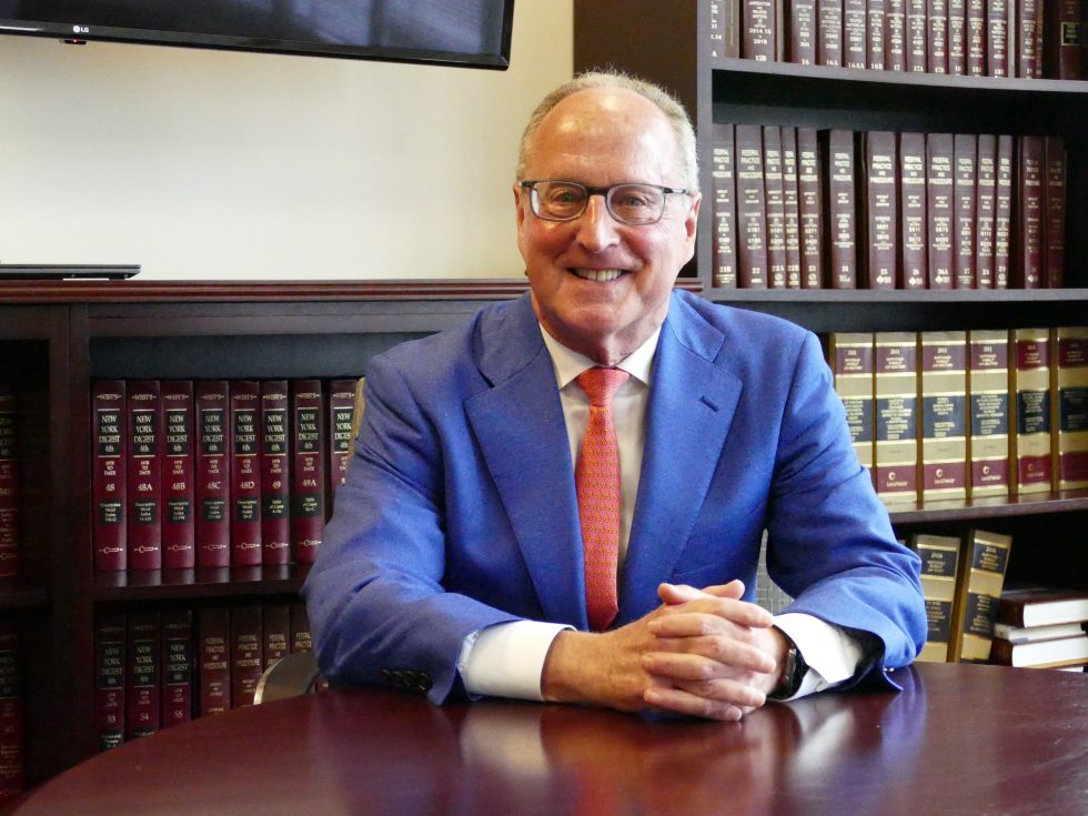 Bern Clair is wearing a blue suit, white shirt and an orange tie. He is sitting at a table and smiling into the camera.