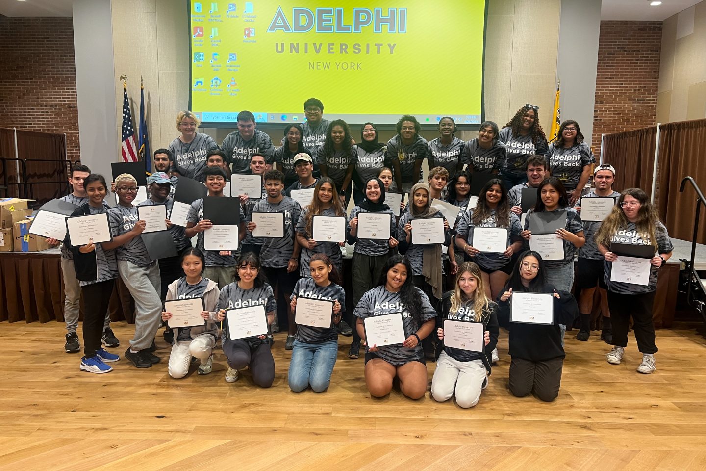 Large group of students wearing tie dye "Adelphi Gives Back" t-shirts hold up certificates. FCAP members on stage behind them.